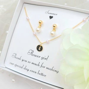 Op de afbeelding: Een witte geschenkdoos met een gouden ketting en oorbellen. De ketting heeft een klein rond hangertje met de letter "E" erop. De oorbellen zijn kleine gouden oorbelletjes met een parel aan elk. De doos bevat een kaart met de tekst "Summer" en "Flower girl" met een bericht waarin de ontvanger wordt bedankt voor het maken van hun speciale dag nog beter.