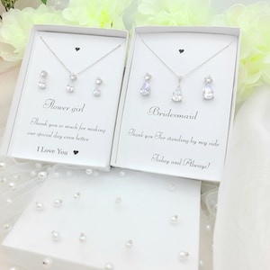 May include: Two white gift boxes containing silver jewellery sets. The box on the left says "Flower girl" and the box on the right says "Bridesmaid". Both boxes have a small heart in the top corner.