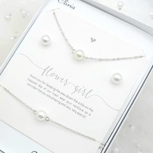 May include: A silver necklace and bracelet set with a single pearl pendant, presented in a white gift box. The set includes two pearl stud earrings. A card reads "flower girl" with a message.