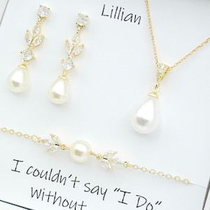 May include: A bridal jewellery set featuring a delicate gold chain necklace, matching earrings, and a bracelet. The necklace and earrings have a pear-shaped pearl pendant with a small leaf design accented with clear cubic zirconia stones. The bracelet has a similar leaf design with a single pearl in the centre. The jewellery is displayed in a white box with the name "Lillian" and the text "I couldn't say "I Do" without".