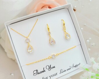 May include: A gold jewelry set in a white gift box, including a necklace, a pair of drop earrings, and a bracelet. Each piece features a clear, pear-shaped or round faceted stone. The box includes the text: Thank You for your Love and Support.