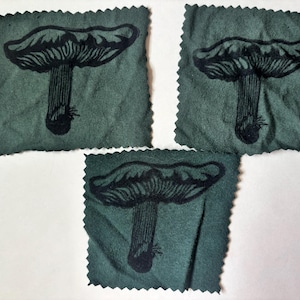 May include: Three square fabric patches in a muted sage green color, each featuring a black ink illustration of a mushroom with a wide cap and visible gills. The edges of each patch are finished with a decorative pinked zigzag cut pattern.