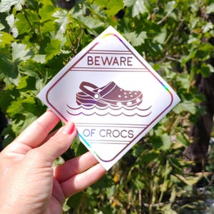 Puede incluir: Una pegatina blanca en forma de diamante con un contorno negro y el texto "BEWARE OF CROCS" en negro. Una ilustración en blanco y negro de un zapato Croc está en el centro de la pegatina, flotando en el agua.