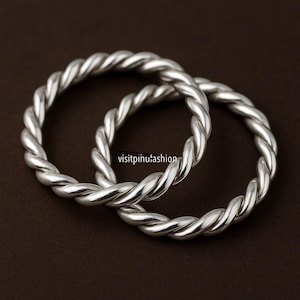 May include: Two silver-colored, twisted metal bangles. The bangles are circular and have a rope-like design. The background is a solid brown color. The text "visitpinhufashion" is visible on the bottom right.