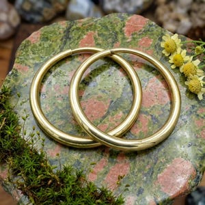 Könnte beinhalten: Ein Paar polierte goldfarbene Creolen mit einer glatten, abgerundeten Oberfläche. Die kreisförmigen Ohrringe sind zusammen positioniert und zeigen ein reflektierendes metallisches Finish sowie ein schlichtes, durchgehendes Design ohne sichtbare Verschlüsse.