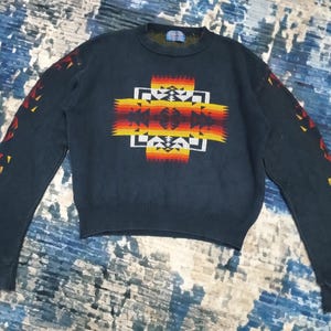 May include: A dark blue sweater with a geometric pattern in shades of red, yellow, and orange. The pattern is reminiscent of Native American art.