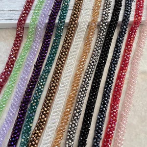 May include: An assortment of beaded trim in various colors, including red, green, purple, gold, silver, and black. The trim is arranged diagonally, showcasing the different colors and textures. The beads appear to be faceted, reflecting light.