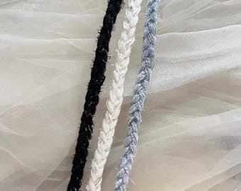 May include: Close-up of three braided cords in black, white, and light blue. Each cord has a textured, slightly fuzzy appearance with metallic silver threads woven throughout. The cords are arranged vertically against a soft, white background.