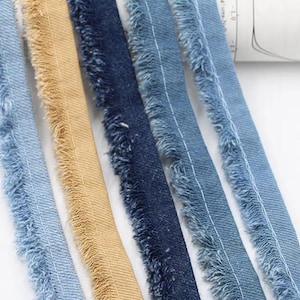 May include: Close-up of several strips of denim ribbon in various shades of blue and tan. The edges are frayed, creating a textured effect. The ribbons are arranged side by side, showcasing the different colours and textures.