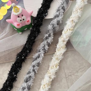 Può includere: Primo piano di tre trecce decorative in nero, grigio e bianco. La treccia nera ha una texture a frange con paillettes. La treccia grigia ha un design intrecciato con una texture sfocata e paillettes. La treccia bianca ha un design intrecciato con una texture sfocata e piccole perle dorate.