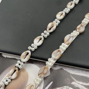 May include: Two beaded seashell and pearl strands. One strand is on a black band, the other on a white band. The seashells are tan and white, and the pearls are small and white. The strands are arranged diagonally.
