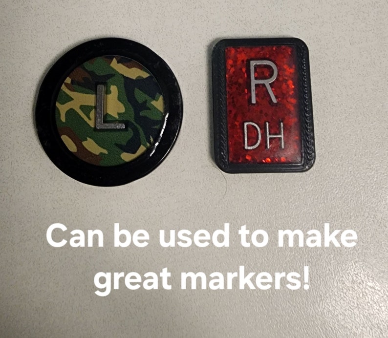Peut inclure: Deux marqueurs ronds avec des bordures noires. Un marqueur a un motif de camouflage avec un "L" blanc au centre. L'autre marqueur est rouge avec des paillettes blanches et les lettres "R DH" au centre. Le texte "Peut &ecirc;tre utilis&eacute; pour faire de grands marqueurs !" est sous les marqueurs.