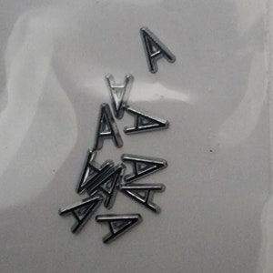 May include: A close-up of a pile of silver letter A charms. The charms are small and have a black outline.