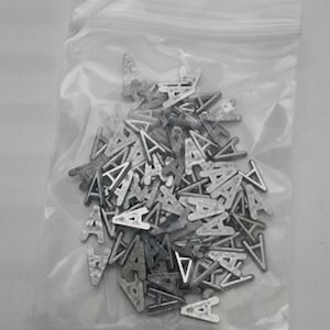 May include: A plastic bag filled with silver metal letter A charms. The charms are all the same size and shape, and they are arranged in a random pattern.