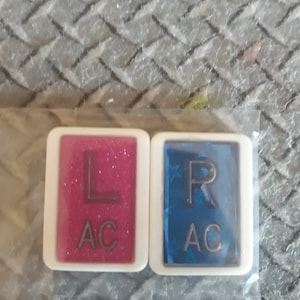 May include: Two rectangular, white-framed accessories, one pink with glitter and the letters "L" and "AC", the other blue with stars and the letters "R" and "AC". The items are in a clear plastic bag.
