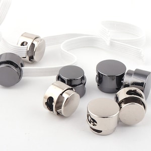 May include: A close-up of eight silver and black metal toggles with two holes each. The toggles are designed to be used with elastic cord or ribbon.