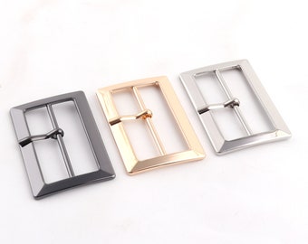 Op de afbeelding: Drie metalen riemgespen in verschillende afwerkingen: zilver, goud en zwart. Elke gesp heeft een rechthoekige vorm met een glad, gepolijst oppervlak.