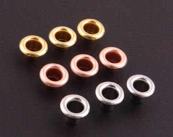 Può includere: Assortimento di otto occhielli metallici in finiture oro, rame e argento. Gli occhielli sono disposti a semicerchio su una superficie nera.