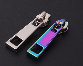May include: Two metal zipper pulls. One is silver, the other is iridescent with a rainbow finish. Both have a rectangular pull tab with a square opening and a curved top. The zipper pulls are on a black background.