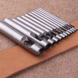 May include: A set of 12 silver metal leather hole punch tools of varying sizes, arranged on a brown leather surface.