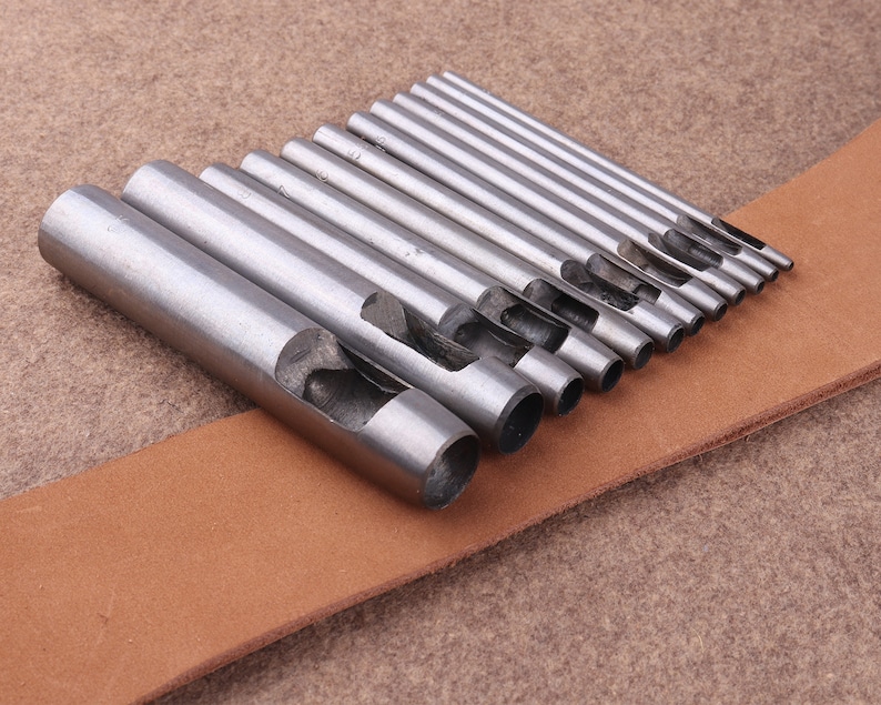 May include: A set of 12 silver metal leather hole punch tools of varying sizes, arranged on a brown leather surface.
