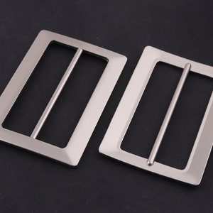 May include: Two silver metal belt buckles with a rectangular shape and a smooth finish. The buckles have a single, horizontal bar across the centre.