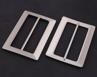 May include: Two silver metal belt buckles with a rectangular shape and a smooth finish. The buckles have a single, horizontal bar across the center.