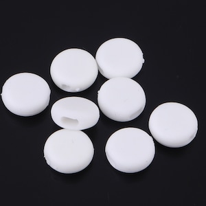 Puede incluir: Ocho cuentas redondas de plástico blanco con un agujero en el centro. Las cuentas están dispuestas en un montón suelto sobre una superficie negra.