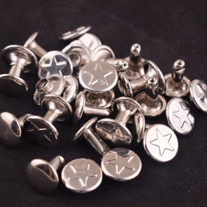 May include: A close-up of a pile of silver metal star-shaped rivets. The rivets have a raised star design and a post for attaching to fabric.