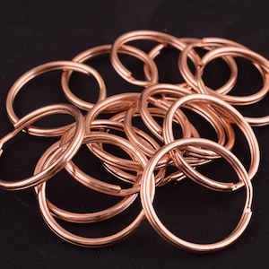 May include: A collection of copper-colored key rings, arranged in a pile against a black background. The key rings are circular, with a split in the ring for attaching keys. The metal has a shiny finish.