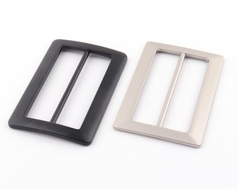Op de afbeelding: Twee metalen riemgespen, een zwarte en een zilveren. Beide gespen zijn rechthoekig van vorm met een enkele, horizontale gleuf in het midden.