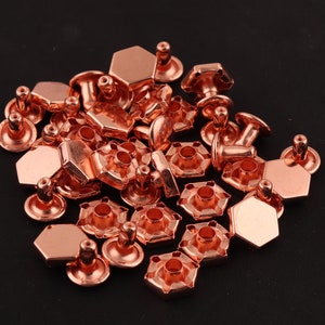 May include: A pile of copper-colored metal rivets with hexagonal heads and posts. The rivets are used for attaching fabric or leather together.