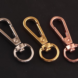 Puede incluir: Tres mosquetones giratorios metálicos en plata, dorado y oro rosa. Cada mosquetón tiene un gancho con resorte y un anillo circular. Los mosquetones se muestran sobre un fondo oscuro, mostrando sus diferentes acabados metálicos.