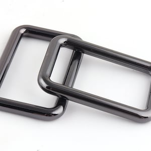 May include: Two black metal D-rings, one slightly larger than the other, are stacked on top of each other. The rings are smooth and have a shiny finish.