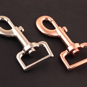 Könnte beinhalten: Zwei Metallschnallen, eine silberfarben und eine roségoldfarben. Beide Schnallen haben einen D-Ring und einen federbelasteten Verschluss.