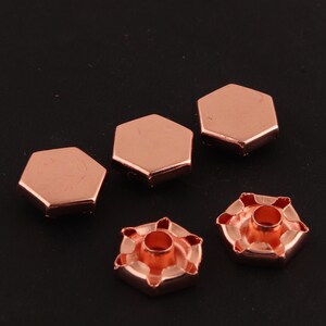 May include: Five rose gold metal hexagonal studs with a hole in the center. Two of the studs are flat and three have a raised center with a hole in the middle.