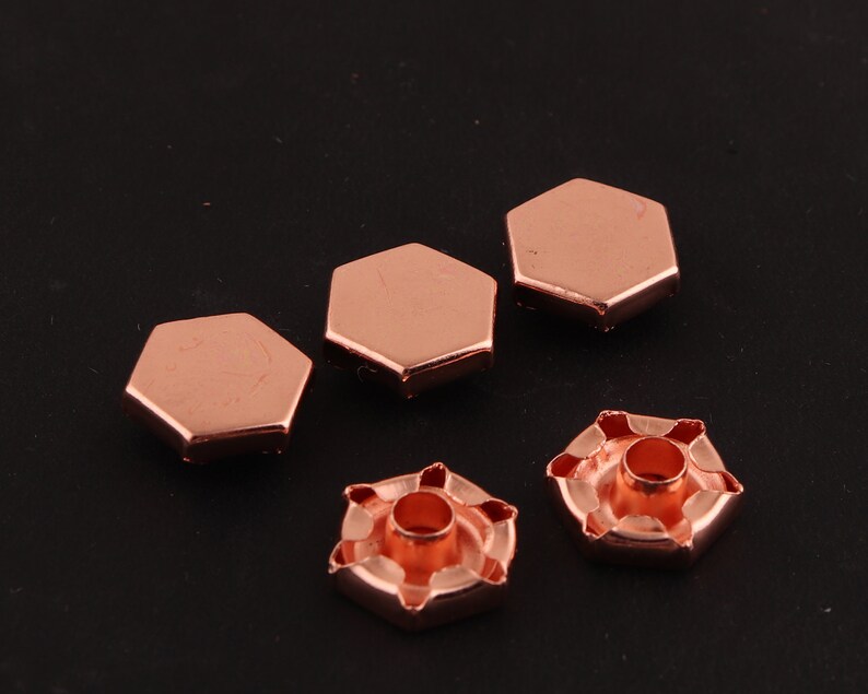 May include: Five rose gold metal hexagonal studs with a hole in the center. Two of the studs are flat and three have a raised center with a hole in the middle.