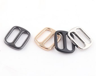 Op de afbeelding: Vier metalen schuifgespen in verschillende afwerkingen: zwart, goud, zilver en gunmetal. De gespen zijn rechthoekig met twee ovale openingen.
