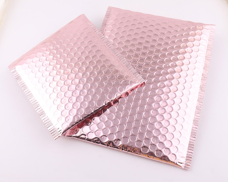 Pu&ograve; includere: Due buste a bolle metalliche rosa, anche note come buste imbottite, sono mostrate su una superficie bianca. Le buste sono realizzate in un materiale sottile e flessibile con uno strato di bolle d'aria per ammortizzare e proteggere.