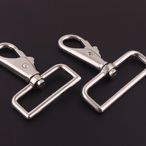 May include: Two silver metal swivel hooks with rectangular loops. The hooks are designed for attaching to straps, belts, or other items.
