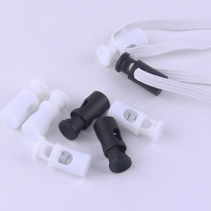 May include: A close-up of black and white plastic cord locks with a spring mechanism. The cord locks are used for adjusting the length of cords or straps.