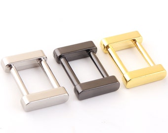 May include: Three metal D-rings in silver, black, and gold. The rings are rectangular with rounded corners and have a smooth finish.