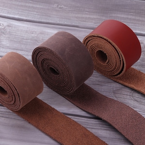 May include: Three leather straps rolled up, one brown, one dark brown, and one red. The straps are on a wooden surface.