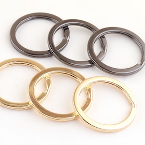 Può includere: Cinque portachiavi, due dorati e tre neri, sono disposti su una superficie bianca. I portachiavi sono tutti circolari e hanno una finitura liscia.