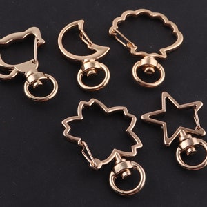 May include: Six gold-tone metal keyrings with different shapes: a cat, a crescent moon, a seashell, a flower, a star, and a circle. Each keyring has a swiveling ring.