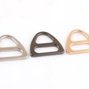 May include: Three metal D-ring buckles in silver, black, and gold finishes. The D-rings are triangular in shape with a rounded edge and a single opening.