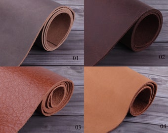 May include: Four different colors of leather rolled up on a wooden surface. The colors are dark brown, burgundy, brown, and tan. The leather is smooth and has a natural grain.