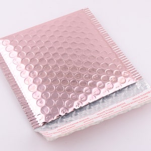 Pu&ograve; includere: Una busta a bolle metallizzata rosa con una striscia adesiva bianca che recita "REMOVE TO EXPOSE ADHESIVE".