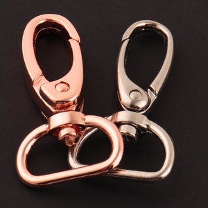 Puede incluir: Dos cierres giratorios de metal, uno en oro rosa y otro en plata. Cada cierre tiene una base de anilla en D y un clip ovalado con resorte. Estos cierres están diseñados para sujetar a bolsos, llaveros u otros accesorios.