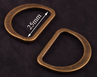 May include: Two antique gold-toned metal D-rings, each with a 25mm inner diameter. The rings are on a black fabric background.